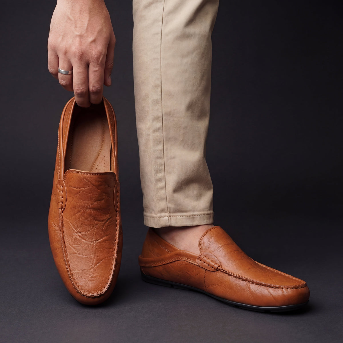 Man holding tan leather moccasin next to foot wearing matching slip-on shoe